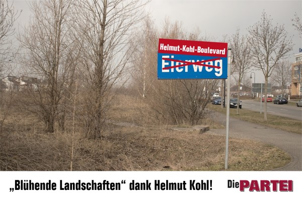 Feld am Eierweg im März. Die Bäume sind kahl, grau und leblos. Das Straßenschild wirkt vergrößert. Die ursprüngliche Bezeichnung Eierweg ist durchgestrichen und wurde durch Helmut-Kohl-Boulevard ersetzt. Unter dem Bild ist der Satz zu lesen: "Blühende Landschaften dank Helmut Kohl!"